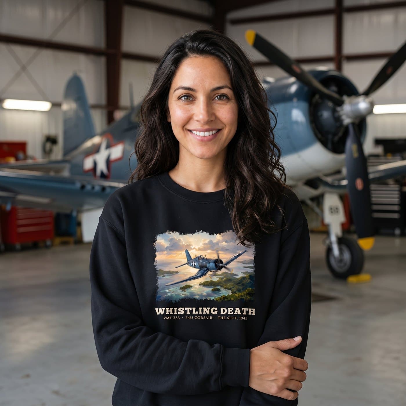 Woman with long dark hair wearing the black Whistling Death crewneck in a restoration hangar with an F4U Corsair
